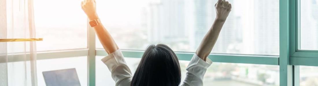 Business achievement concept with happy businesswoman relaxing in office or hotel room, resting and raising fists with ambition looking forward to city building urban scene through glass window